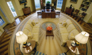 Overhead shot of the Oval Office featuring the Presidential seal in the center of the room and Resolute desk in the background.