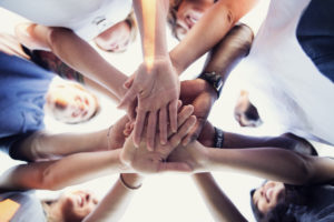 A diverse group of young adults placing the hands in the middle of a huddle to show unity.